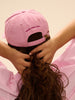 Person wearing a pink cap and dress, adjusting hair against a beige background